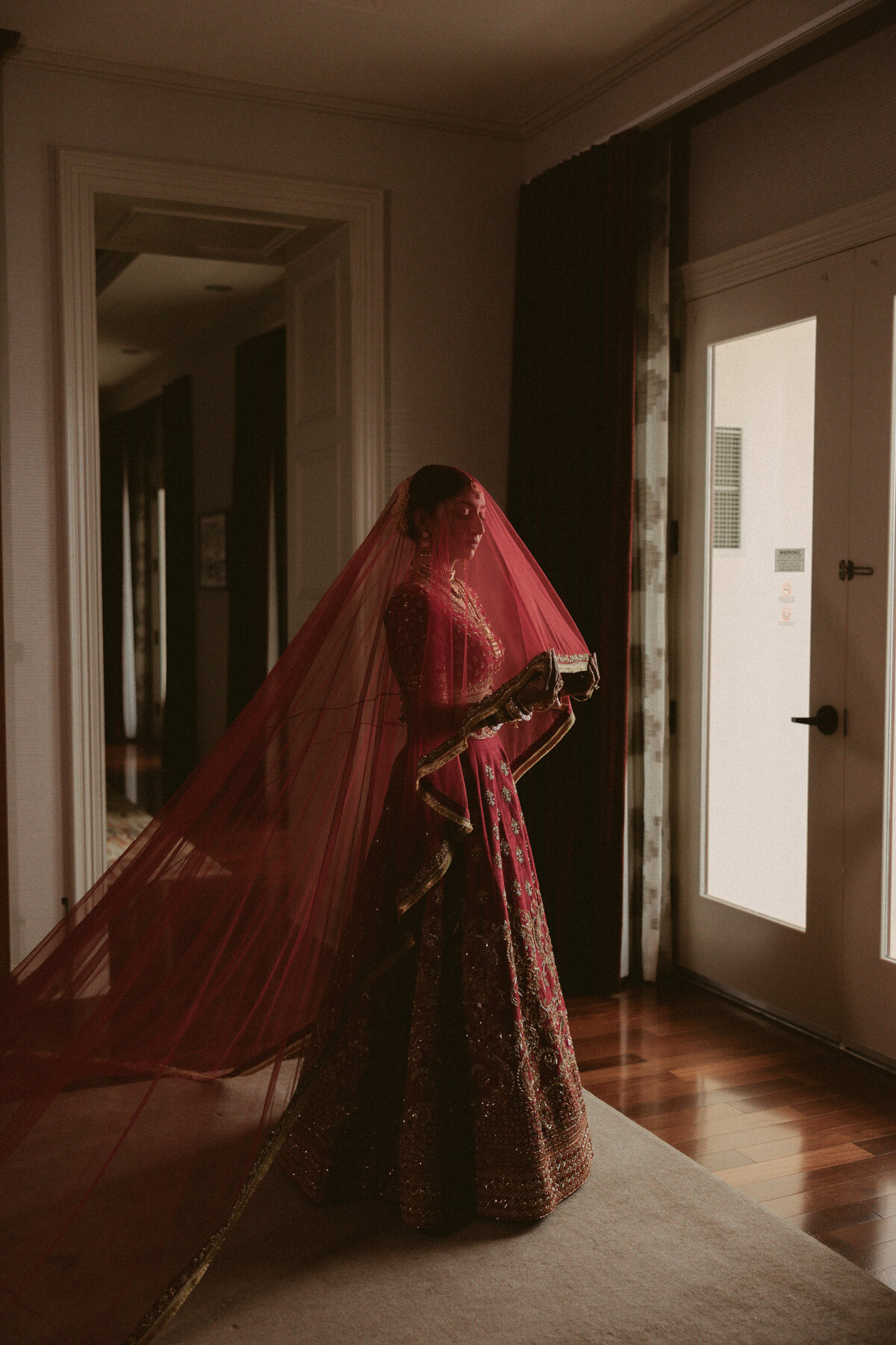 South Asian bride wearing a red bridal lehenga with a long veil, standing in soft indoor light before her ceremony.