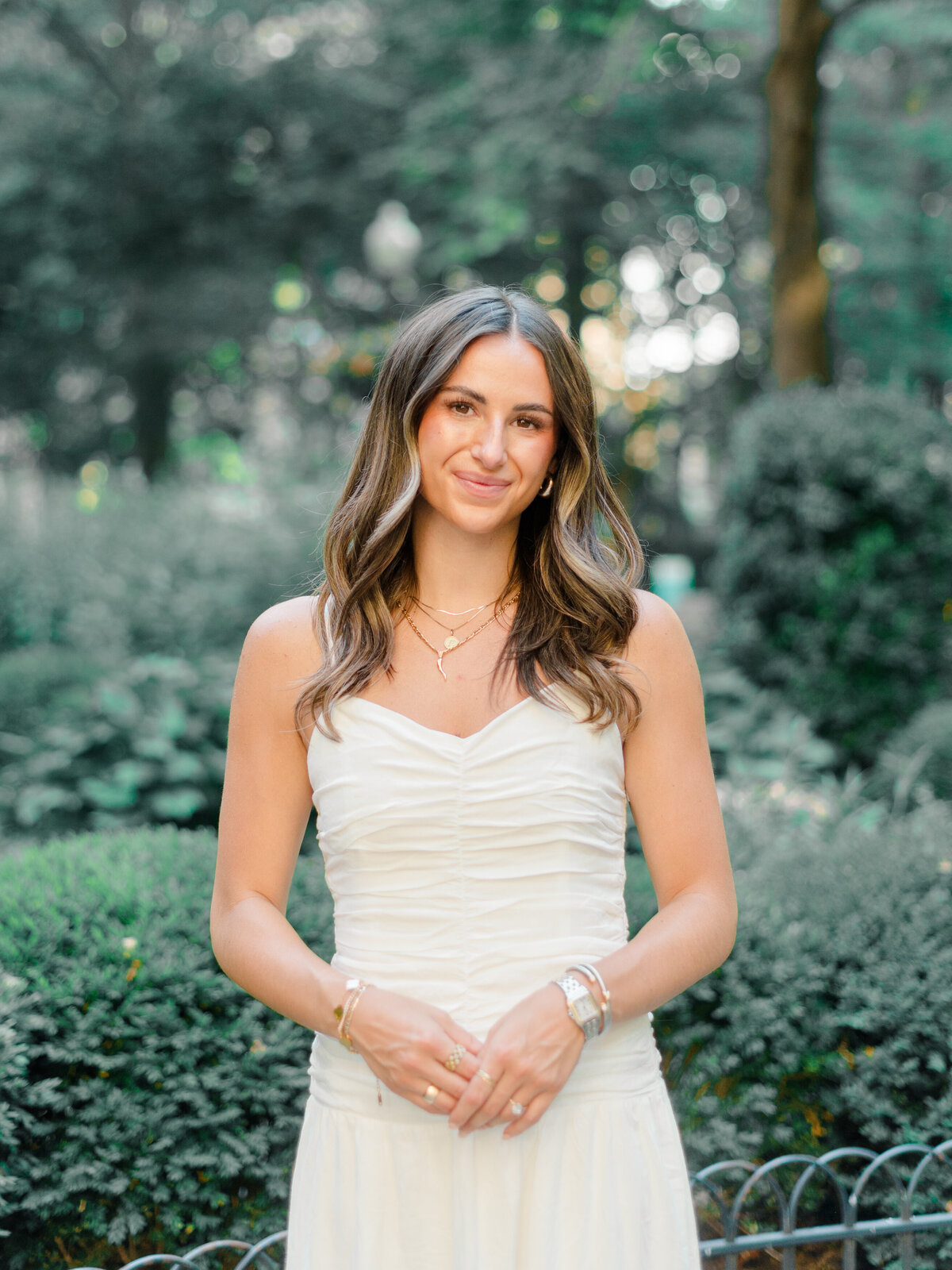 A bride-to-be stands gracefully in Rittenhouse Square, wearing a ruched white dress and layered gold jewelry. Soft greenery and dappled light frame her serene expression in this timeless portrait, captured by Elizabeth Kane Photography, a Luxury Philadelphia Wedding Photographer.