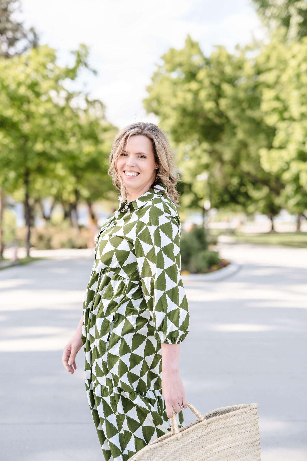 Female physician looking over shoulder outdoors in green patterned dress holding woven straw bag in Kelowna.