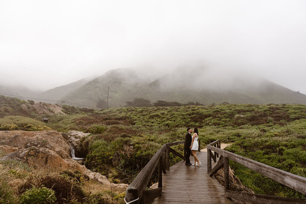 A couple stands on a wooden bridge in a foggy, green landscape, sharing a quiet moment together.