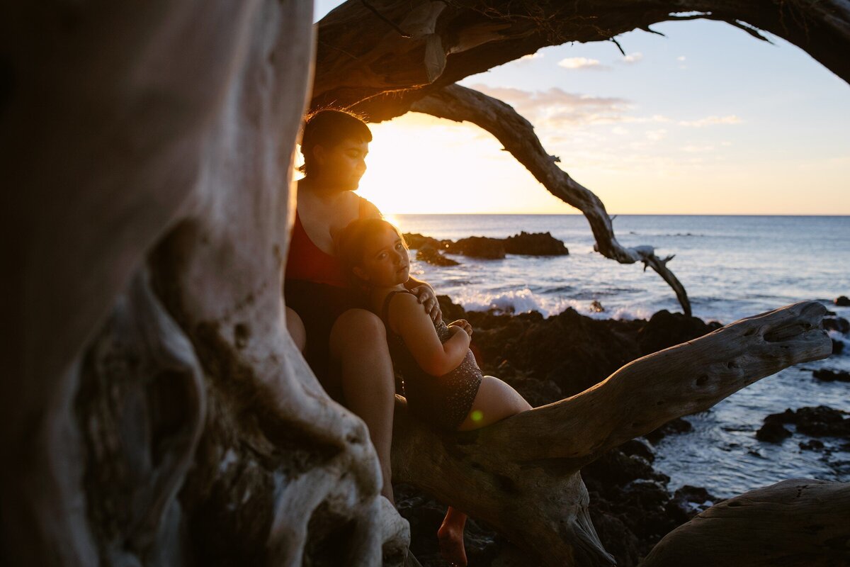 mom and daughter  watching sunset on the rocky kohala coastline from a driftwood tree