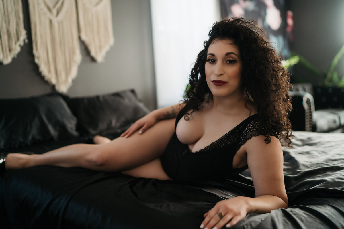Curly brown haired woman posing on a black bed while leaning on her elbow  in a luxury boudoir photo session