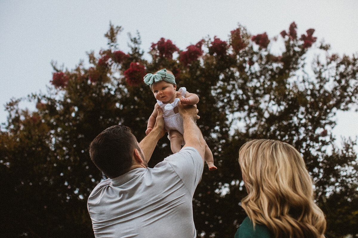 mom and dad hold daughter above heads with trees and flowers in background 