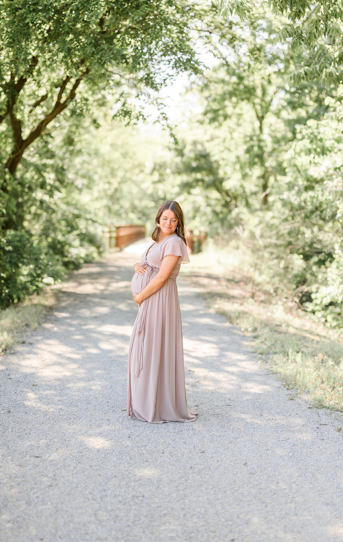 Pregnant woman standing on a tree-lined trail in a soft mauve dress during a golden-hour maternity session in Kansas City.