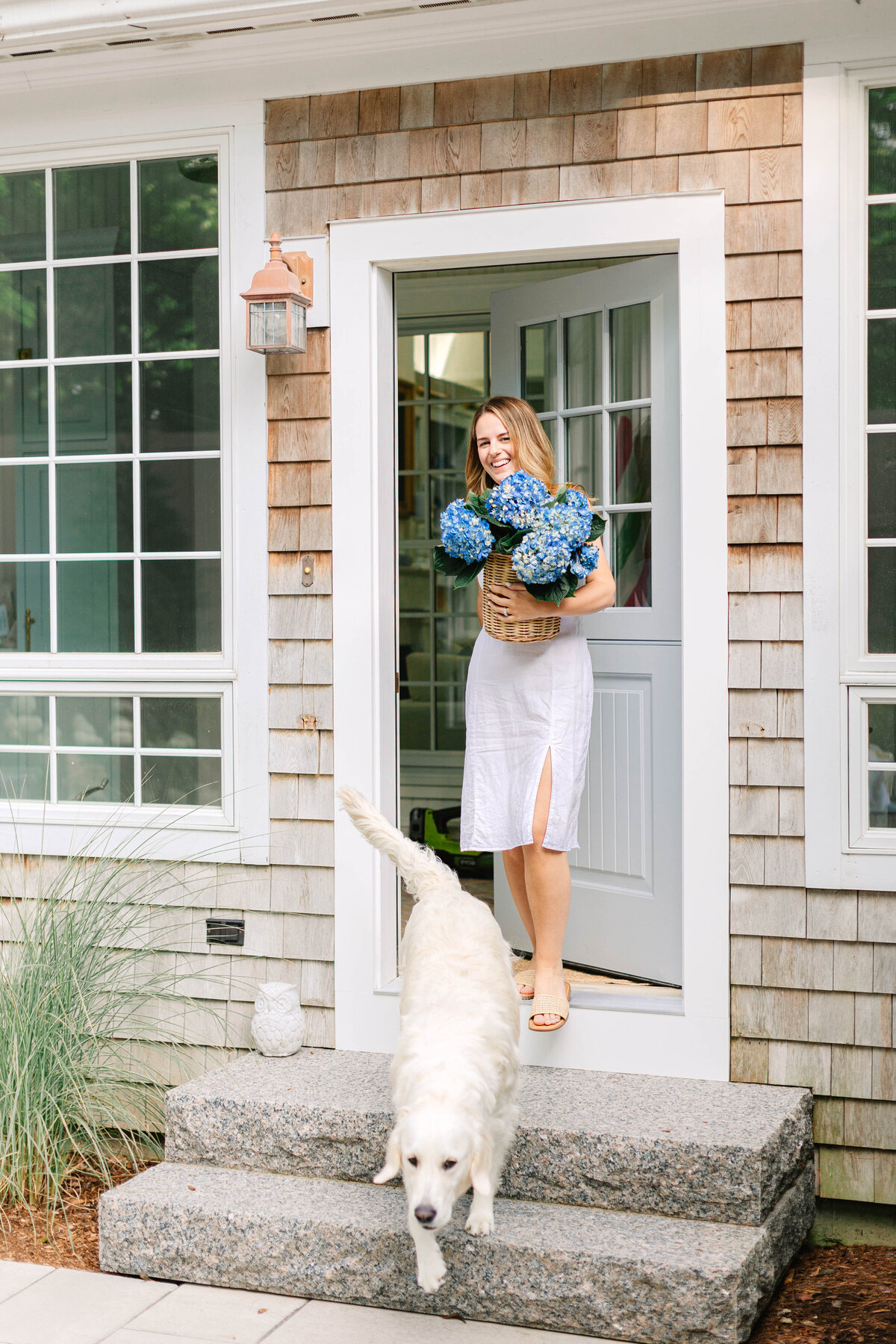 A smiling woman in a white dress steps out of a coastal-style home, holding a wicker basket filled with blue hydrangeas, while a fluffy golden retriever trots down the stone steps.
