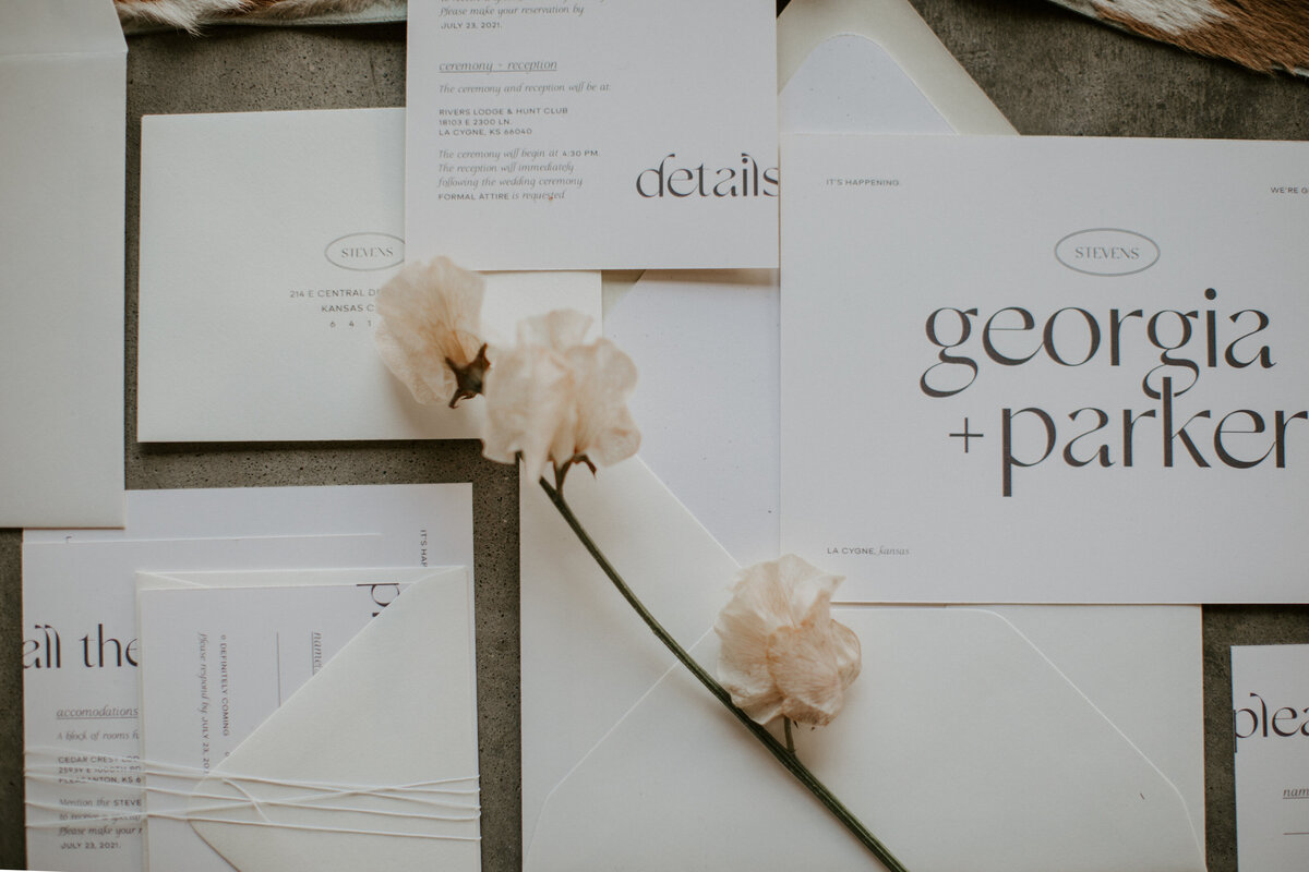 A flower atop various white wedding stationery with brown font.