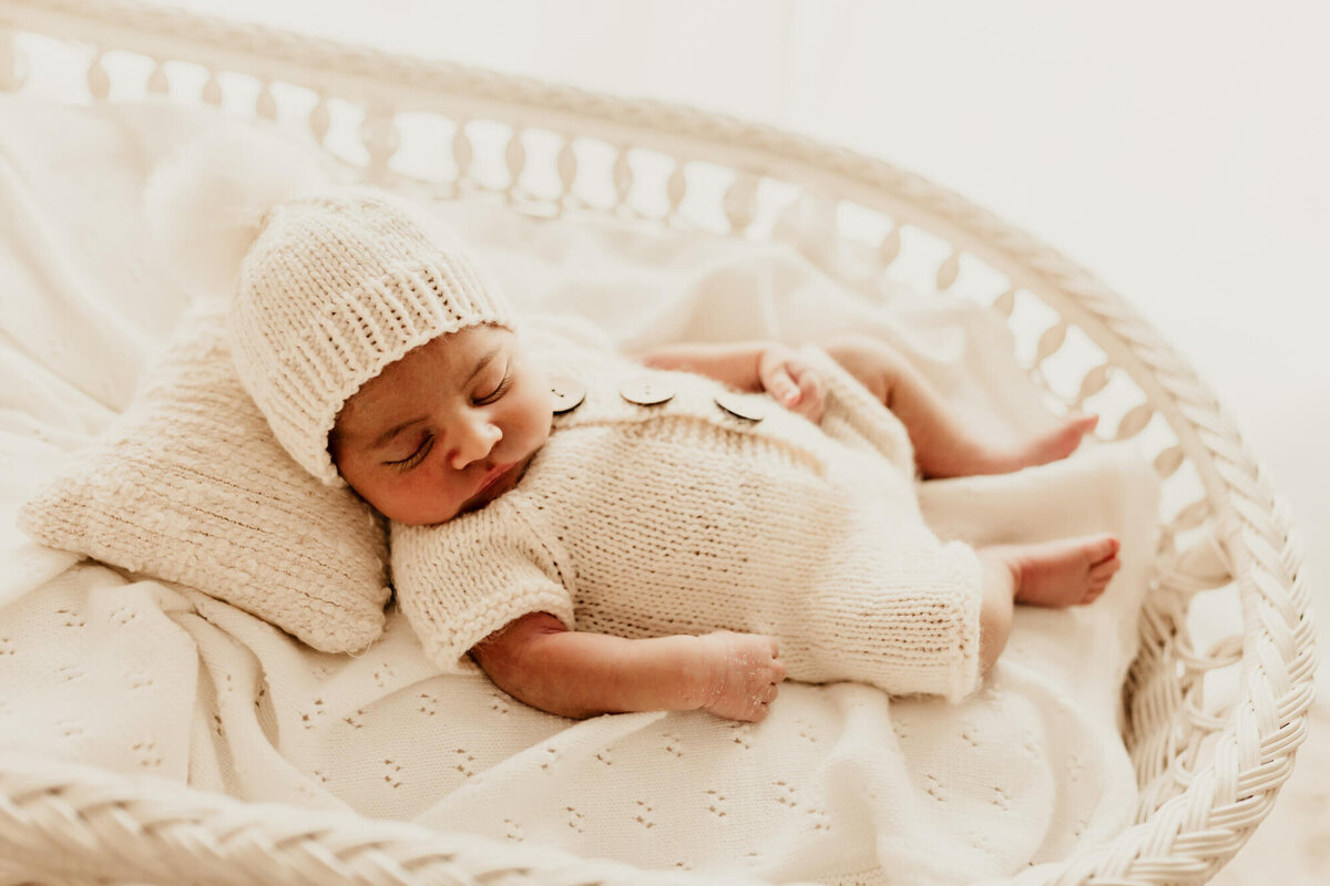 Baby boy wearing a white romper and beanie sleeps on a white blanket.