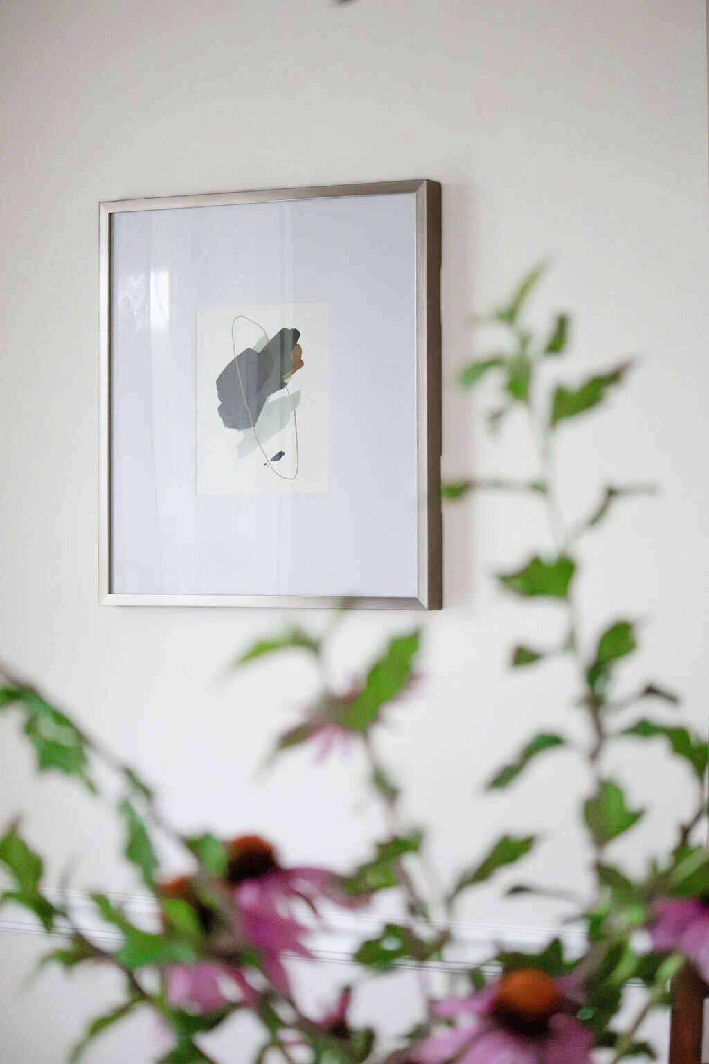 A framed square abstract artwork with muted tones of gray, green, and terracotta hangs on a light beige wall. The blurry foreground shows the stems and purple flowers of coneflowers, suggesting they are in a vase nearby but out of focus. The lighting is soft and natural.