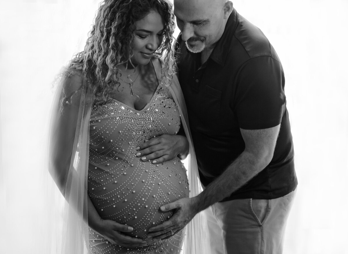 Expectant mom and dad pose for maternity photos. Mom is wearing a long beaded gown with one hand under and one hand atop of her belly. Dad is standing sideways with forehand underneath her belly. Mom and dad are looking down and smiling at her bump.