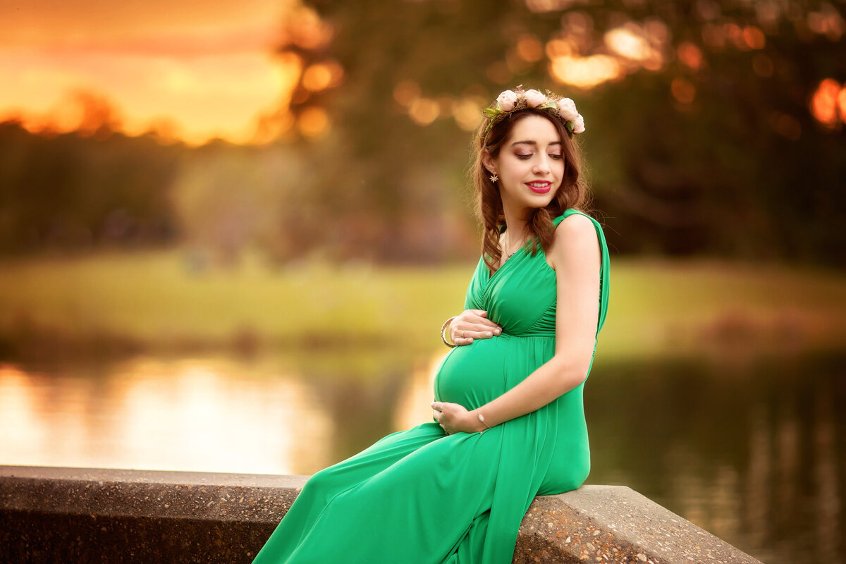 Petite pregnant woman is sitting on a bench in City Park during sunset.  She is holding her belly.  She has a green sleeveless dress, dark hair, and a flower crown.  Her eyes are looking over her shoulder and she has a soft smile.