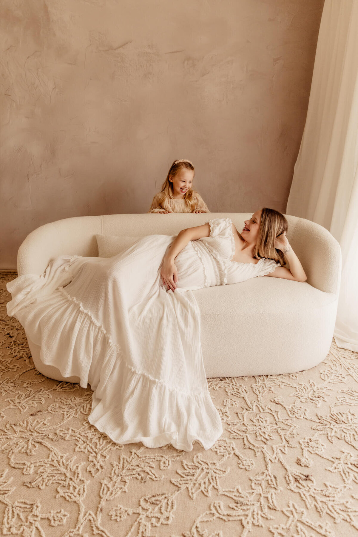 Girl plays peek-a-boo with her pregnant mother who is laying on a white sofa.