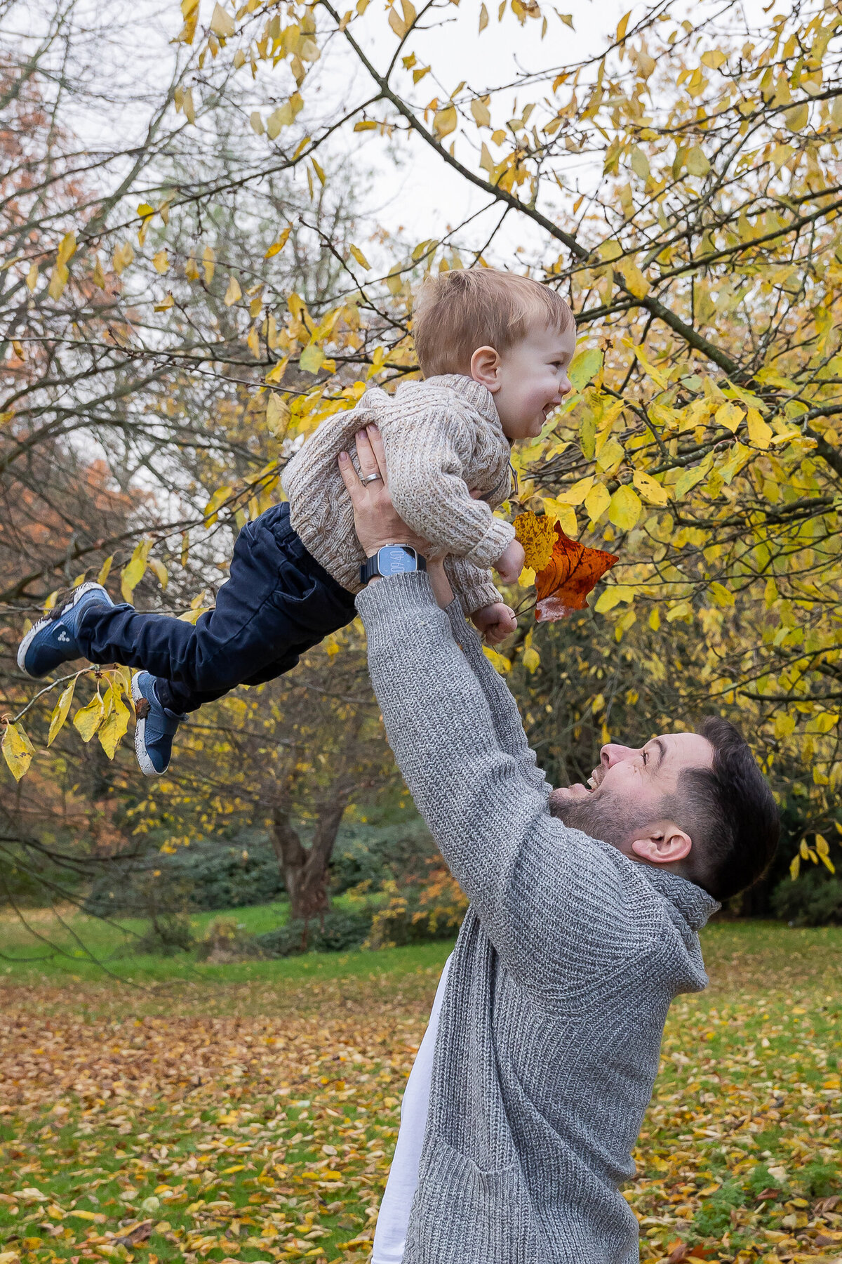 A man lifting a baby high in the air under a tree with yellow autumn leaves.