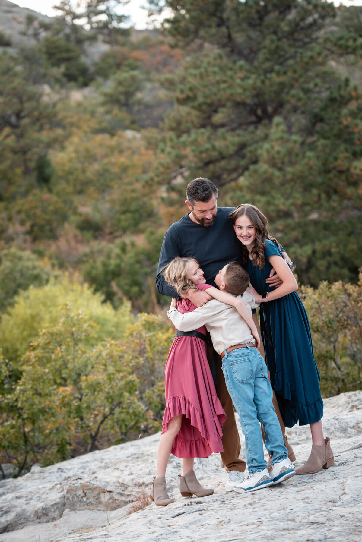 A father in a blue sweater hugs his three children while standing on a mountain trail