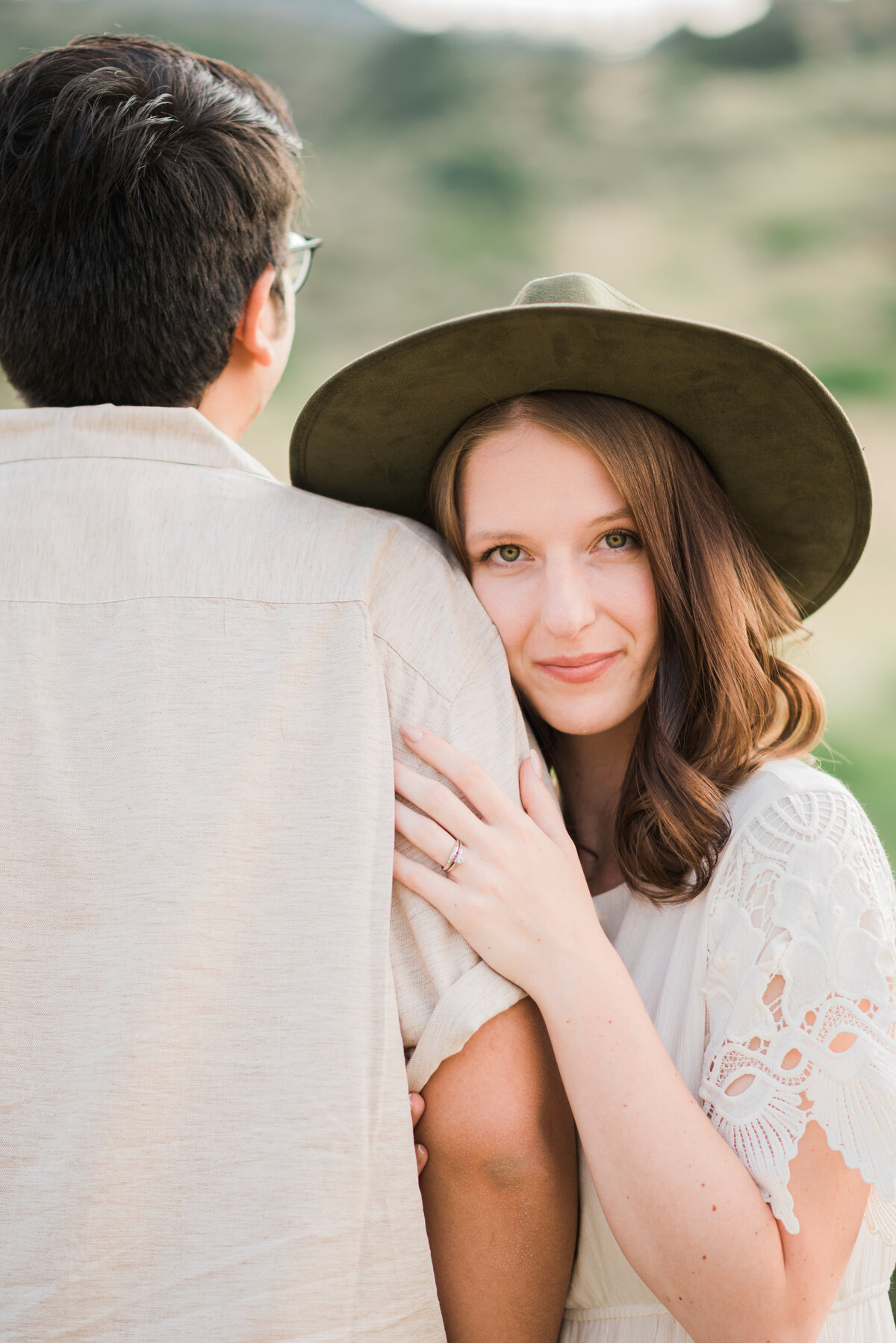 woman holding her fiances arm while looking over his shoulder and showing off her engagement ring while wearing a white blouse and wide brimmed hat