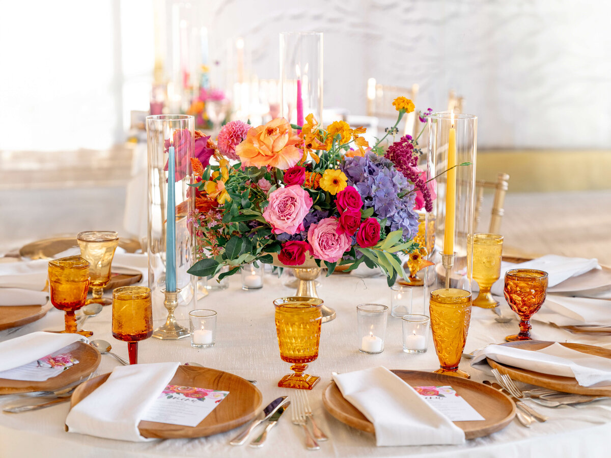 A beautifully set table with a floral centerpiece featuring colorful roses, hydrangeas, and other flowers. Amber glasses and gold-trimmed dinnerware are neatly arranged, with white napkins and candles completing the elegant setting.