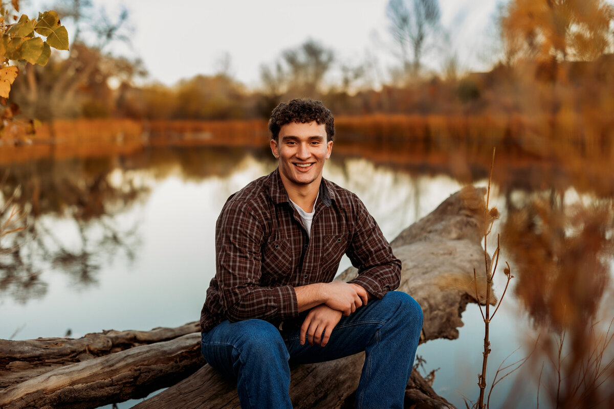 Senior boy wearing a plaid button up sits on a log over hanging a lake on a fall day during his high school senior photos