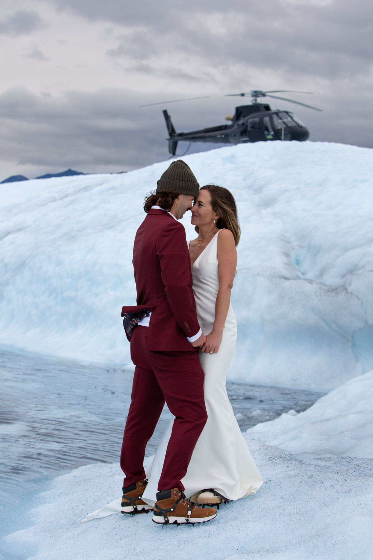 A bride and groom stand on a glacier with a helicopter parked behind them.
