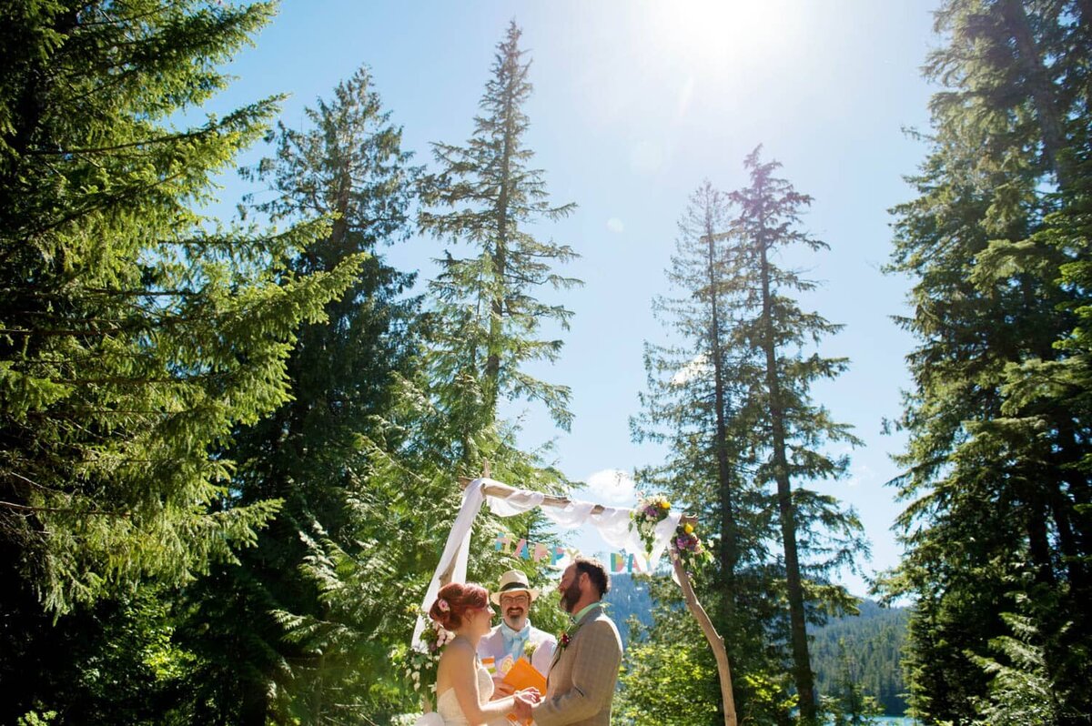 a couple stand underneath tall trees and their wedding arbor that says happy day