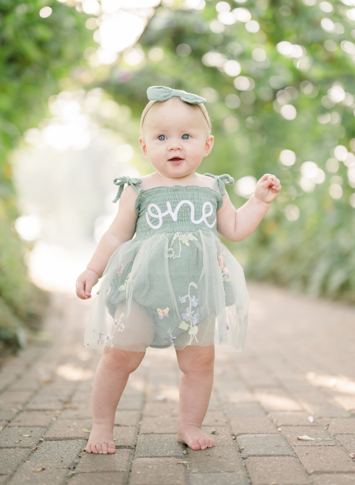 Toddler standing on a brick path outdoors, smiling with hydrangeas in soft focus in the background.