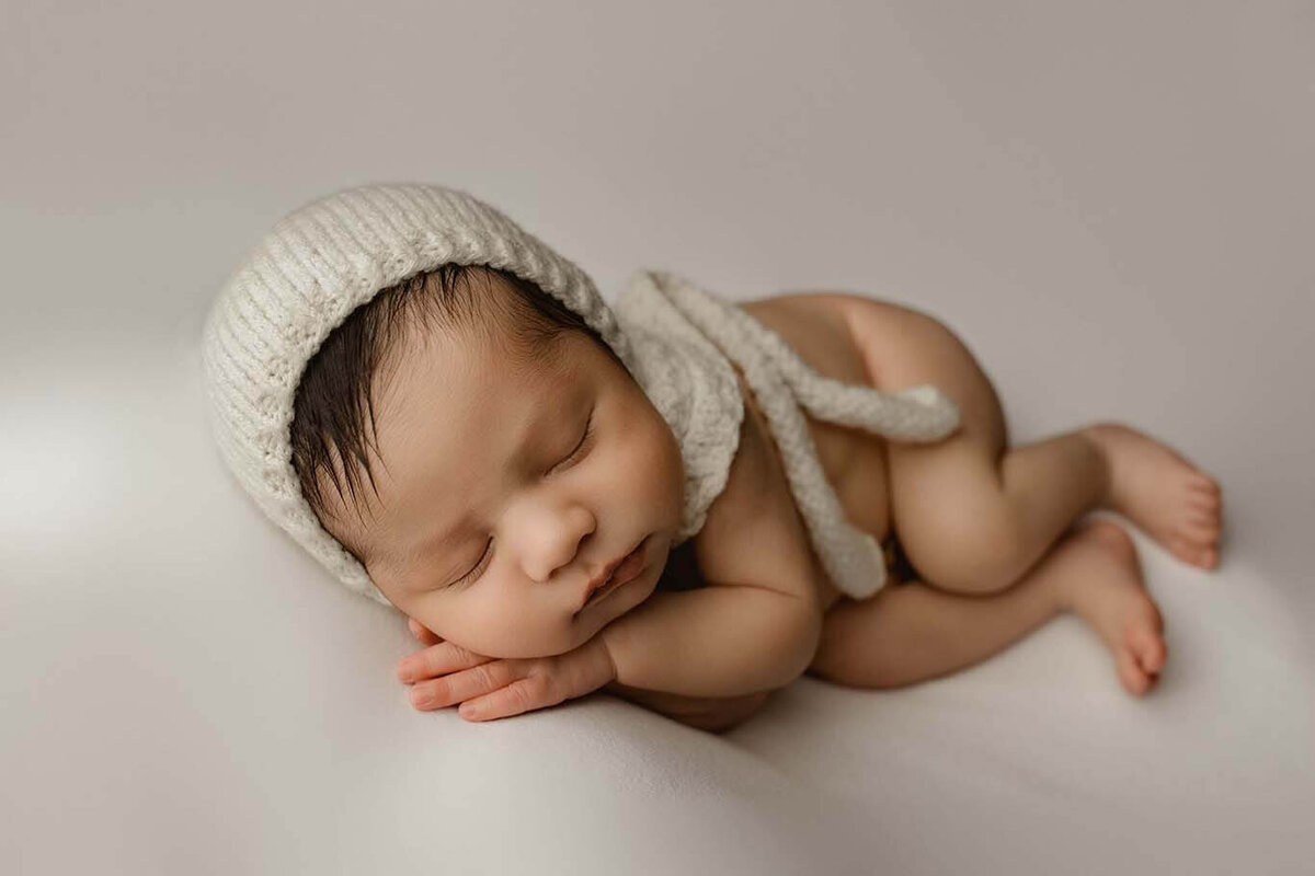 A newborn boy asleep on his side with his hand under his cheek wearing a white bonnet on a white backdrop