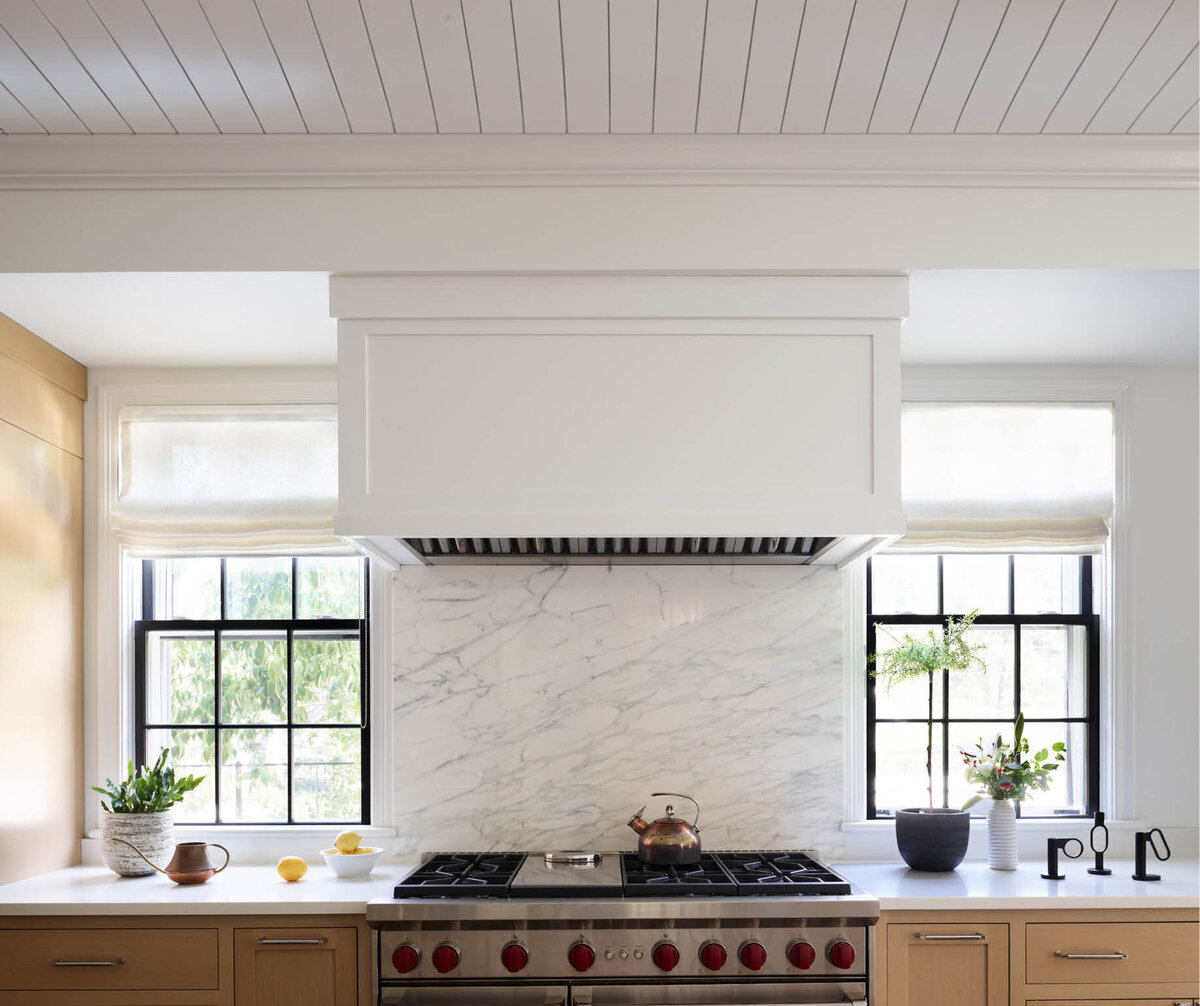 Admire the sophisticated kitchen in a historic Hingham home, featuring a custom hood integrated with the soffit, a slab marble backsplash, and a perfectly positioned range between original windows.