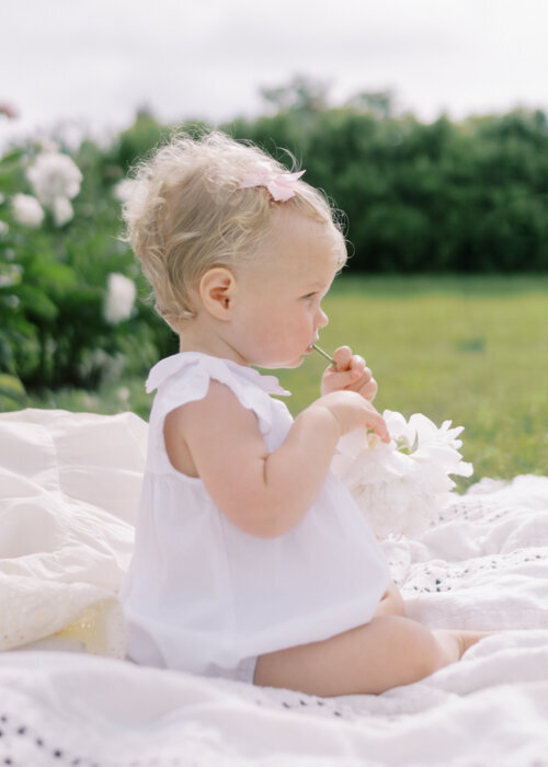 A one year old girl dressed in a white bubble chews on the end of a Peony flower during her Evanston newborn photographer session