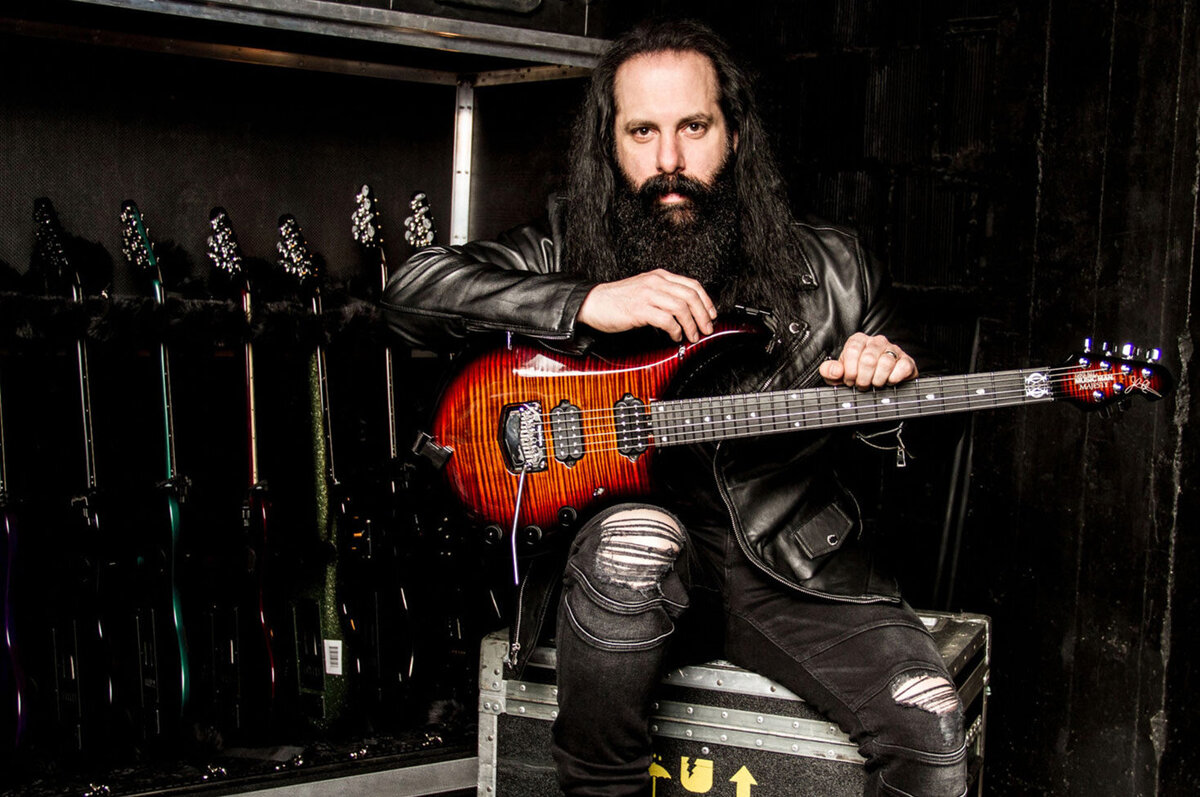 John Petrucci musician portrait  seated on gear case next to rack of guitars guitar across his lap