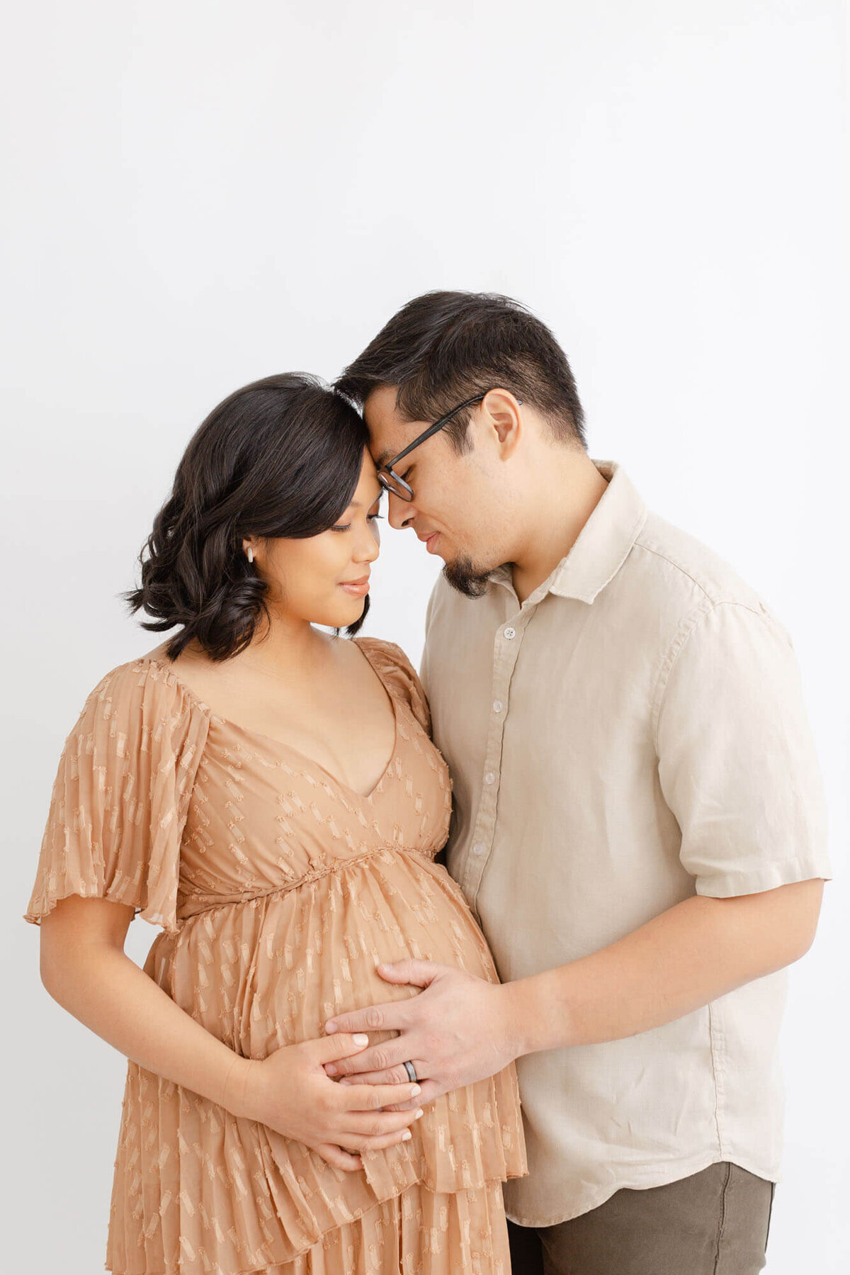 Woman in a light brown dress holding her baby bump standing next to her husband. Their foreheads are touching and they are holding her belly together.