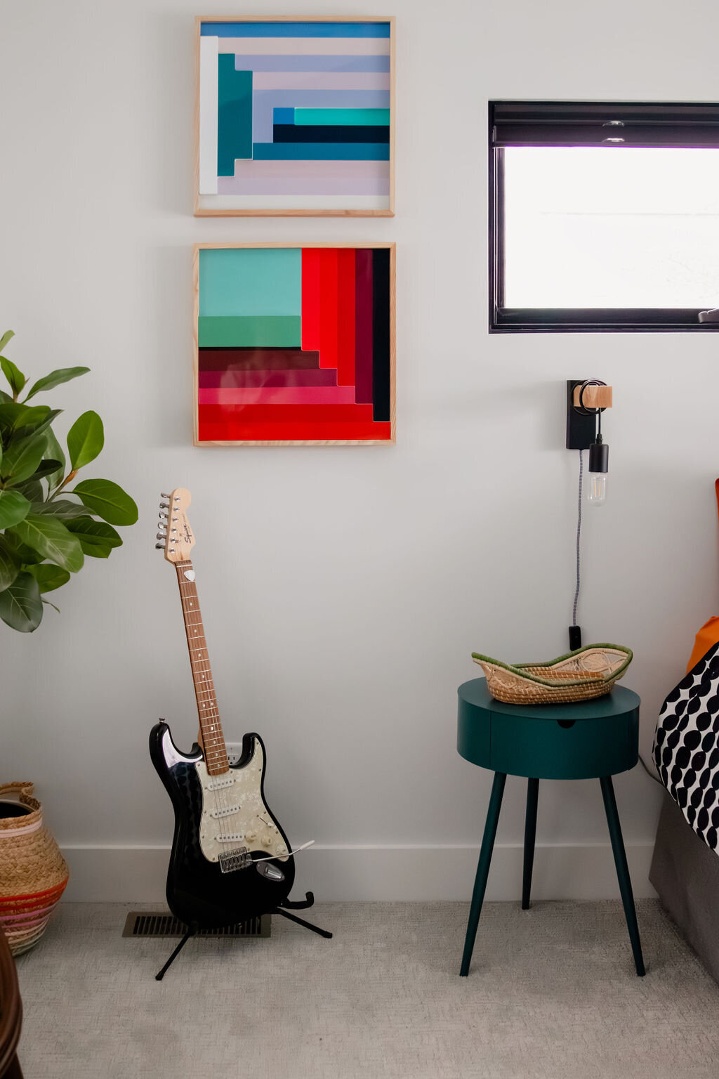A modern bedroom with light gray walls features a black electric guitar, possibly a Fender Stratocaster or Squier Stratocaster, leaning against the wall on a black stand. Above the guitar, two framed abstract artworks with geometric shapes in shades of blue, teal, and red are hung. To the right, a small round teal side table with black metal legs holds a woven basket. The edge of a bed with black and white patterned bedding and orange sheets is visible. A small black-framed window is above the bed, and a fiddle leaf fig plant in a woven basket is to the left of the guitar. A black adjustable wall lamp with a wooden accent is mounted near the bed. The floor is covered in light gray carpet.