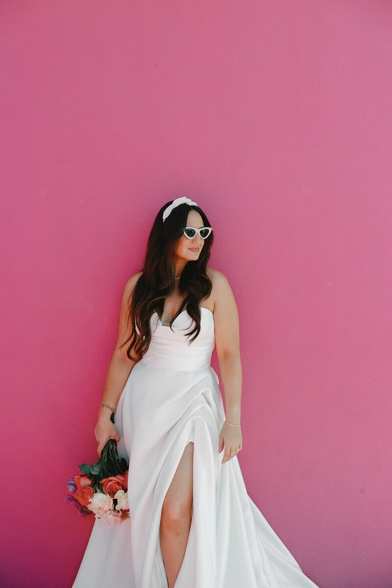 bride wearing sunglasses and holding bouquet posing in front of pink wall at penthouse at riverside wharf​