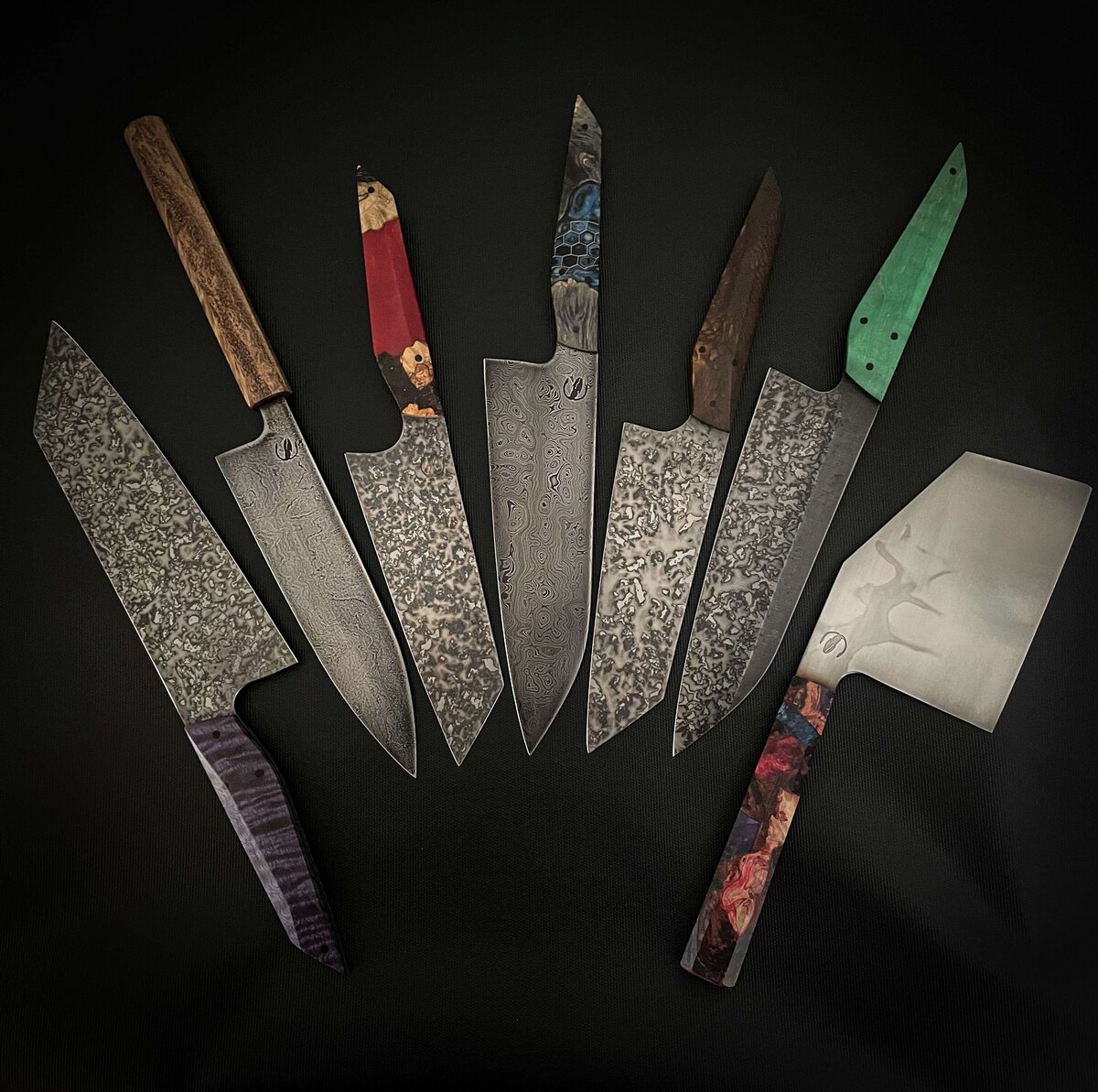 seven handmade chefs knives lay next to each other with multi coloured handles