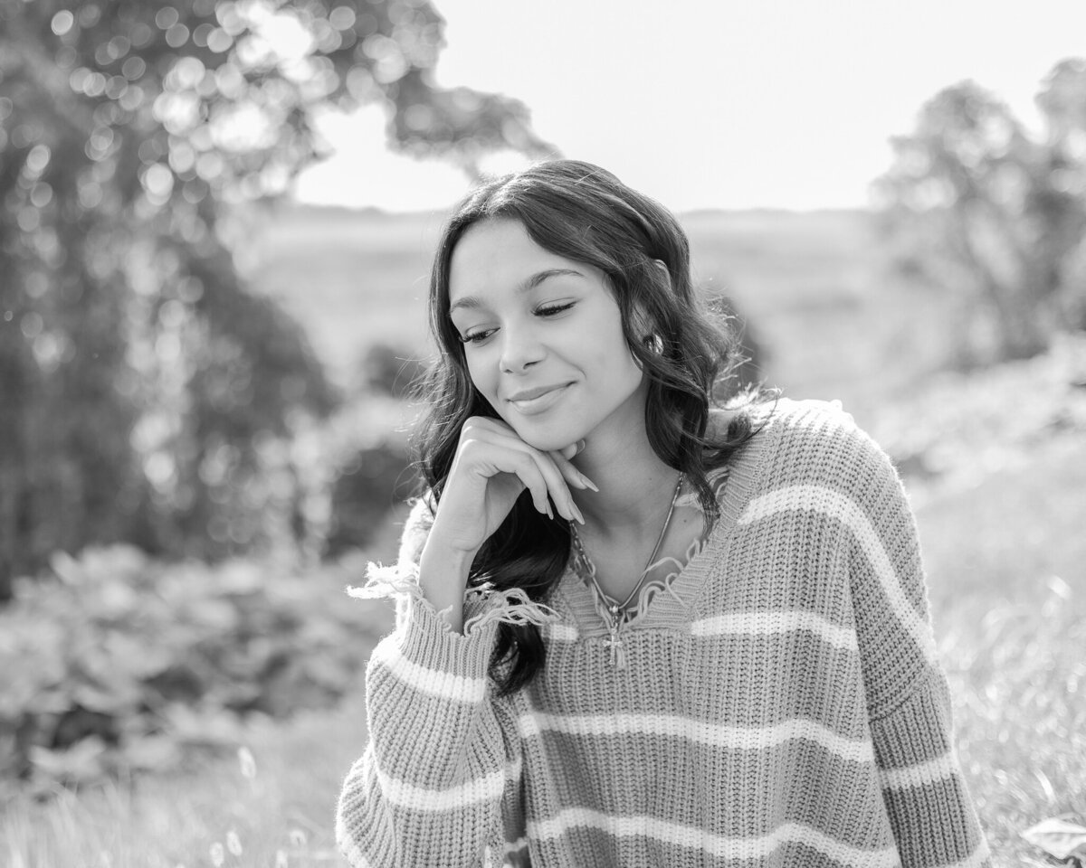 black and white photo of a female high school senior, with long dark hair, sitting in grass with trees behind her, hand under her chin, looking down, smiling softly