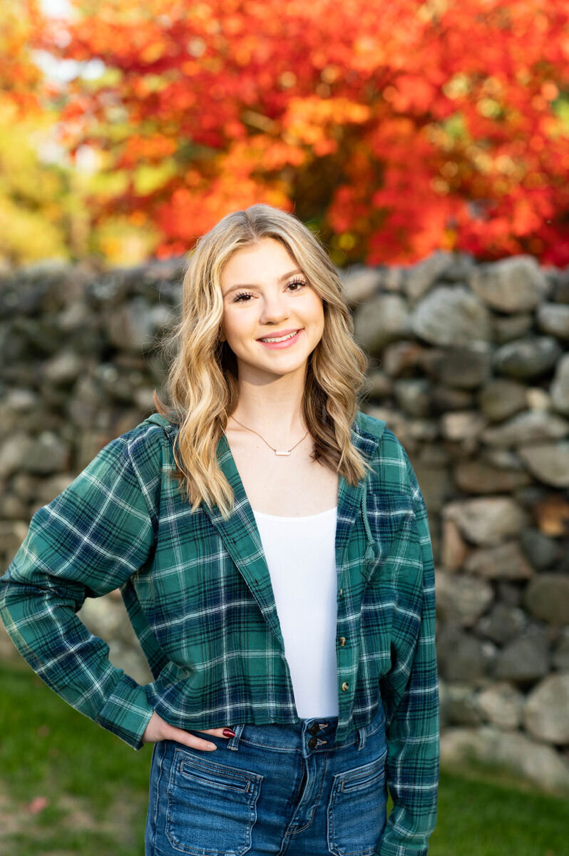 A girl in a flannel shirt with her hand on her hip and fall colored trees behind her.