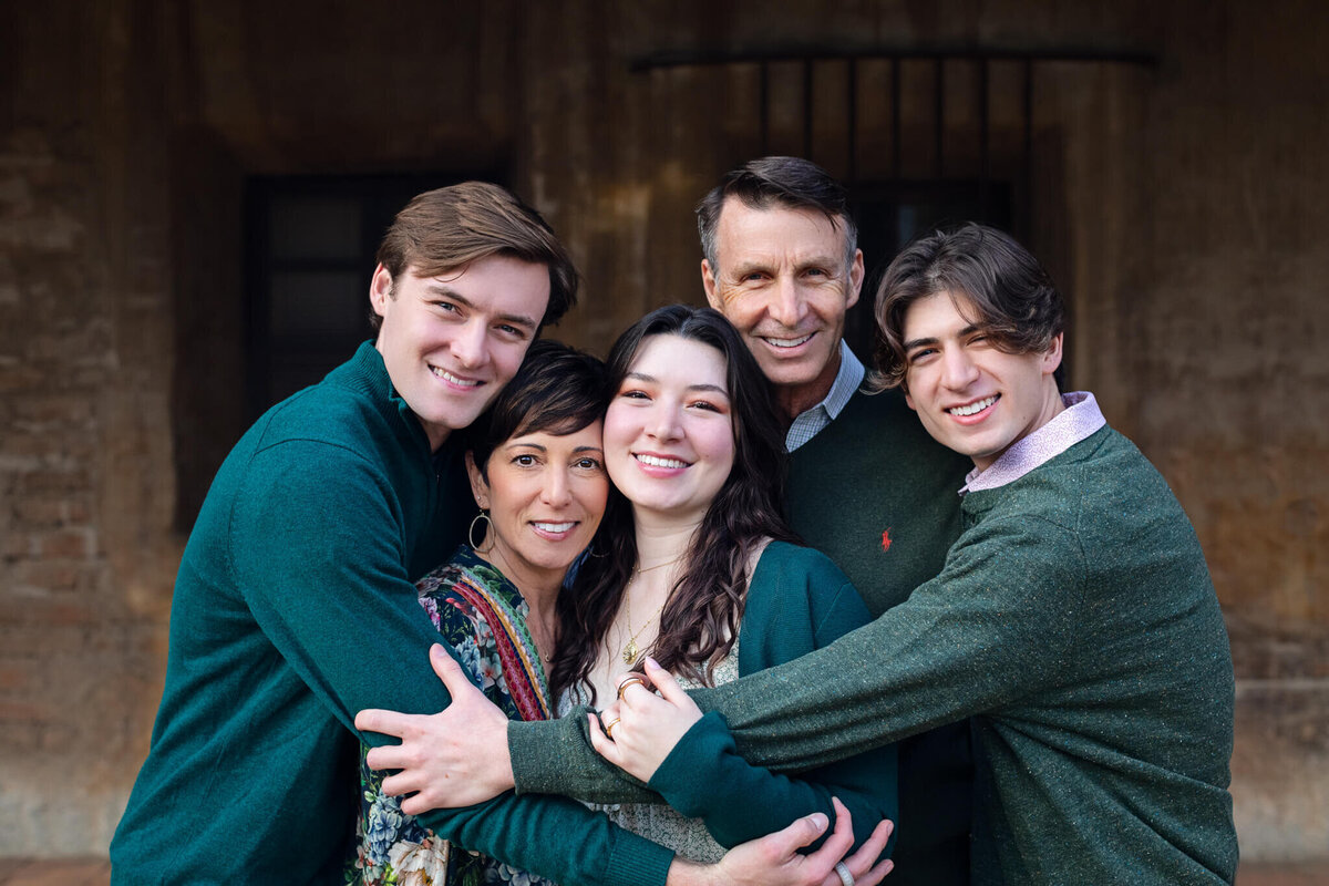 A family of five huddling together in a hug while smiling at the camera, wearing shades of green at Mission San Juan Capistrano.