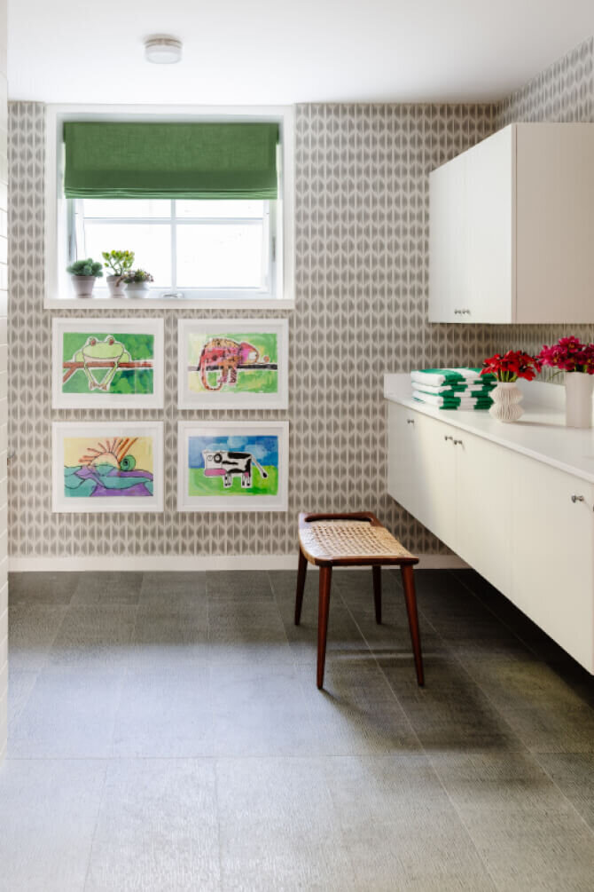 Kid-friendly bathroom design features grey tile, a white vanity, Schumacher wallpaper, colorful art, and green window shades for a playful yet stylish space.