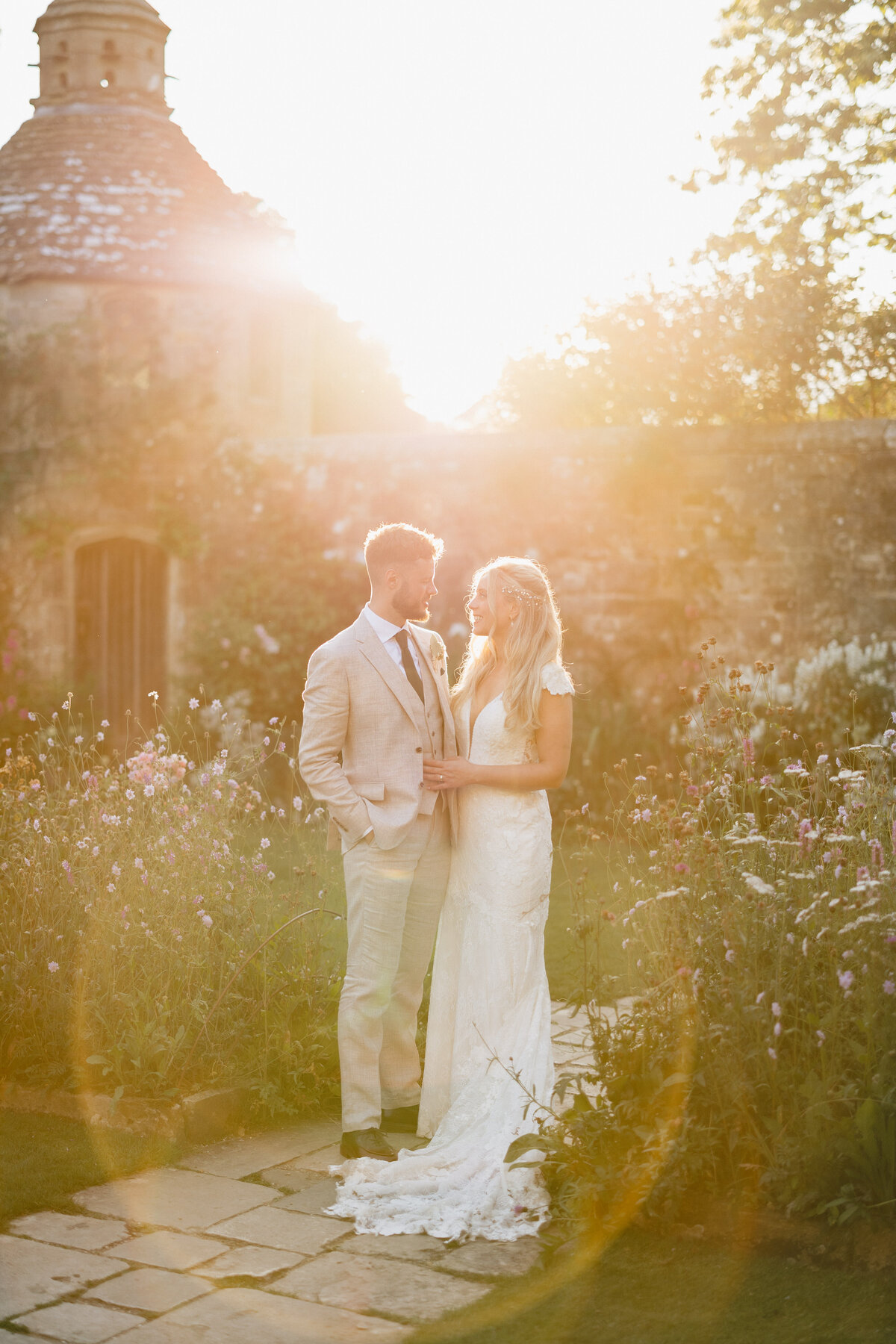 A couple stands on a garden path at Nymans National Trust during sunset. The man is in a light beige suit, and the woman wears a white lace gown. They are surrounded by flowers, with a stone building in the background, bathed in warm, golden light.