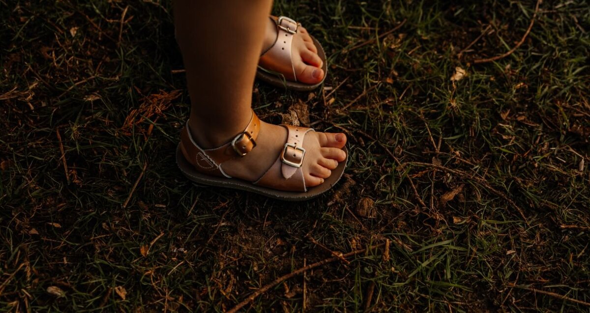 A person's hand adjusts the gold buckle of a Salt Water Sandals Surfer style sandal on their foot, while seated on a wooden surface. Close-up footwear shot