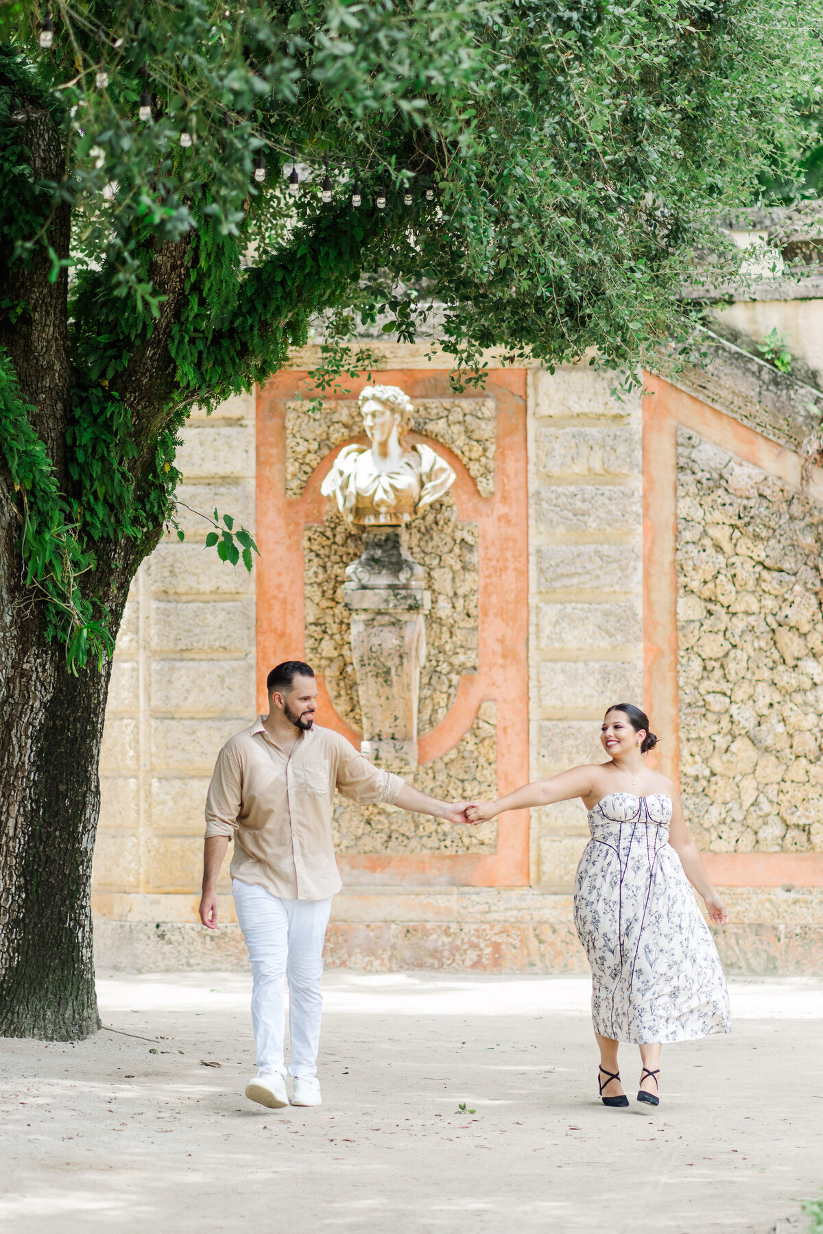a couple holding hands while walking towards  the camera at the vizcaya museum in miami