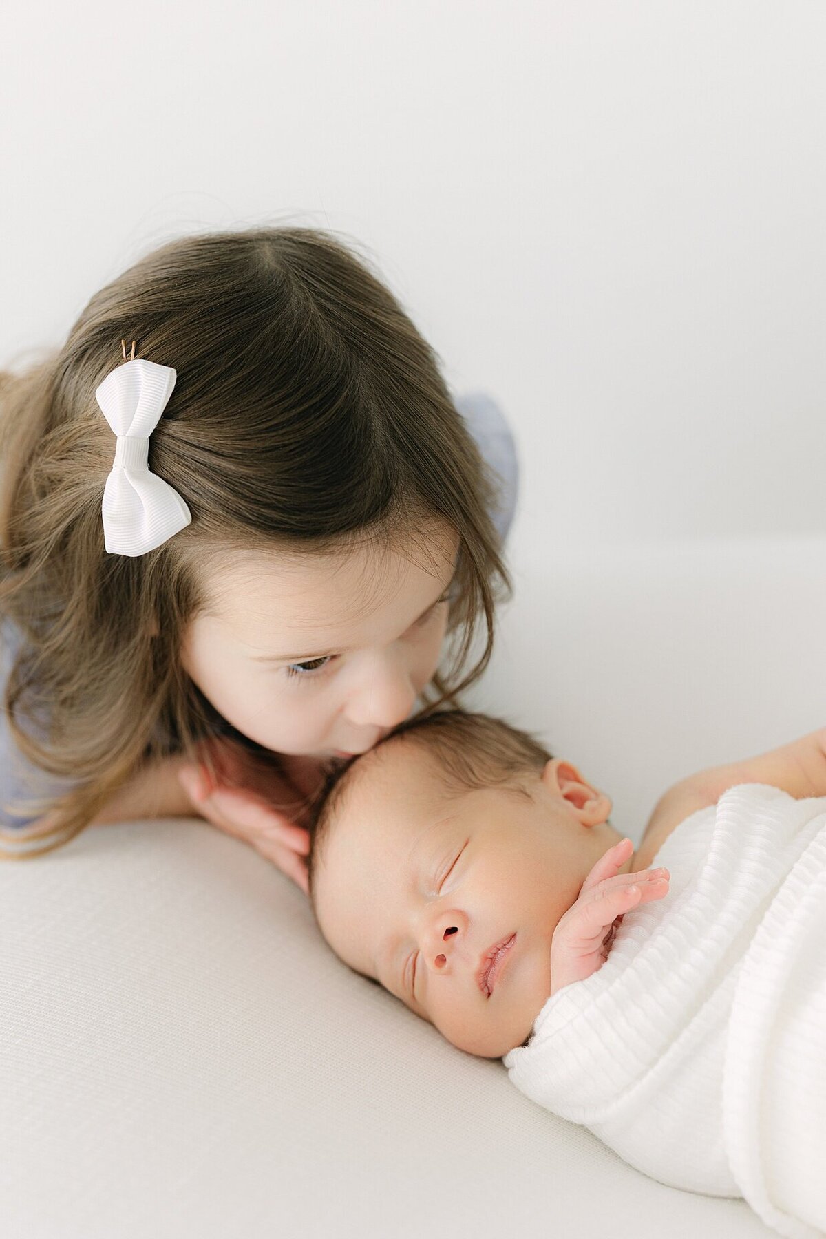 Toddler girl with a white bow in her hair kisses her newborn brothers head as he sleeps swaddled in white with Indianapolis newborn photographer Brittney Lear