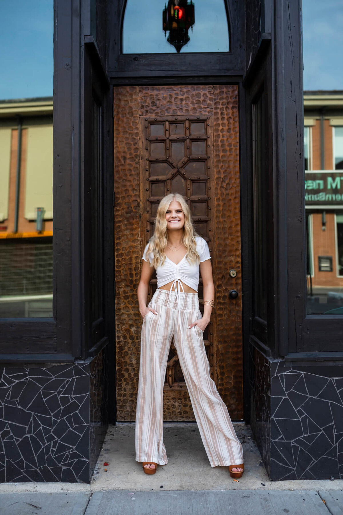 Confidently posed beautiful blond high school senior girl wearing a white crop top and white striped palazzo pants standing in a downtown intricate doorway. Captured by Springfield, MO senior photographer Dynae Levingston.