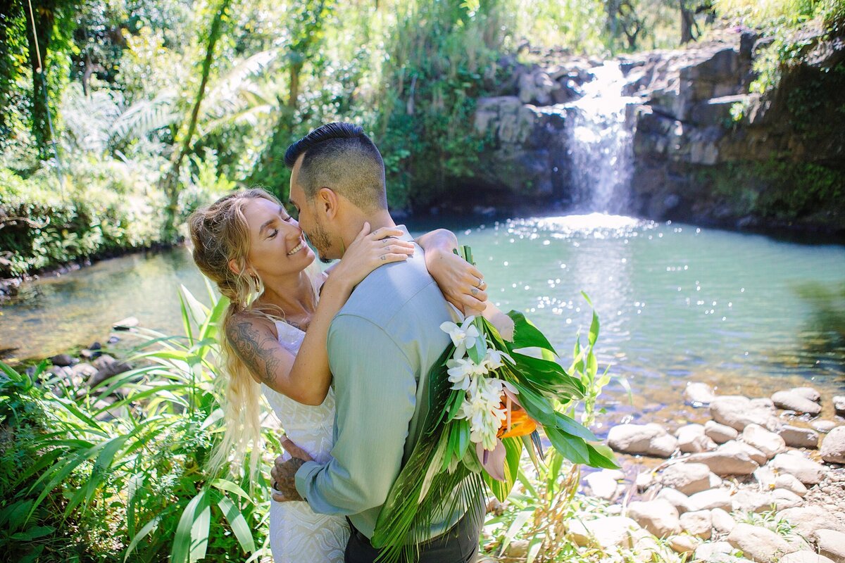 A bride and groom stand in front of a waterfall for their Hawaii elopement