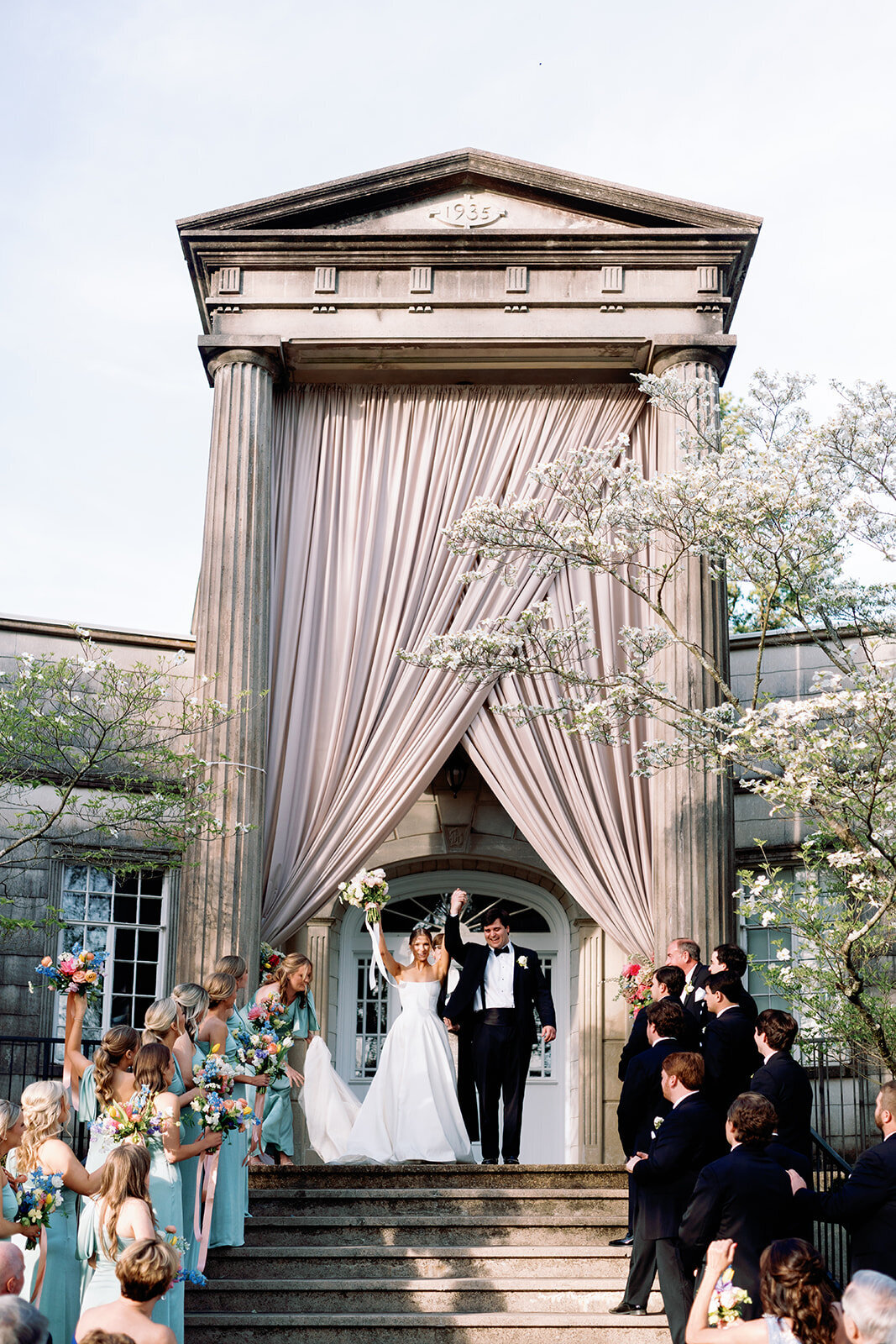 Bride and groom raising their hands in celebration after being pronounced married during their ceremony in front of the grand columned entrance at Burritt on the Mountain in Huntsville, Alabama, photographed by Perry Hancock.