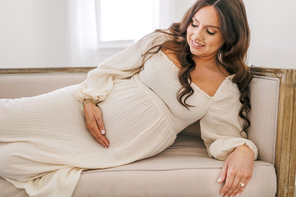 A pregnant mama in a white dress laying with her hand on her belly.