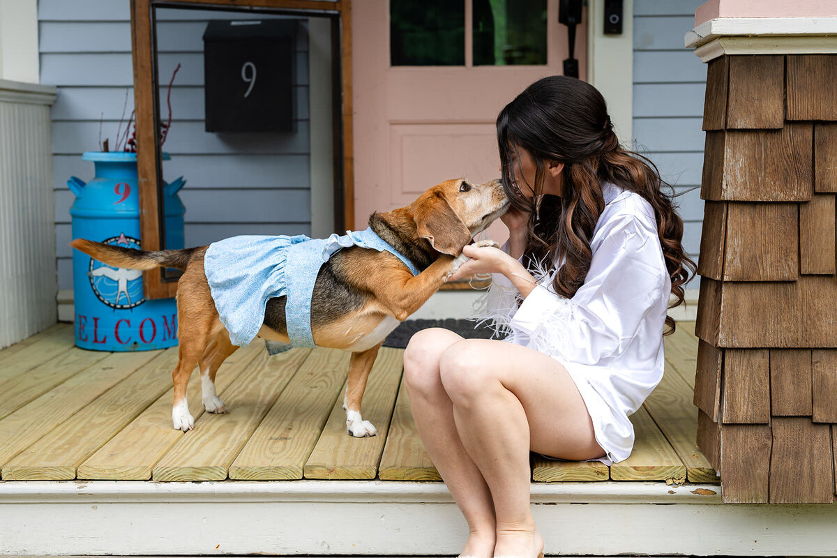 A bride sitting on a porch step, lovingly interacting with a beagle wearing a blue dress, in front of a 'Welcome' bucket and a pink house.