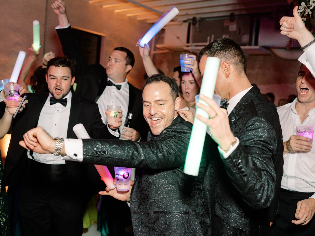 Guests revel in a vibrant party atmosphere, pumping their arms with illuminated sticks, showcasing excitement and energy as they dance and enjoy the lively music throughout the wedding.