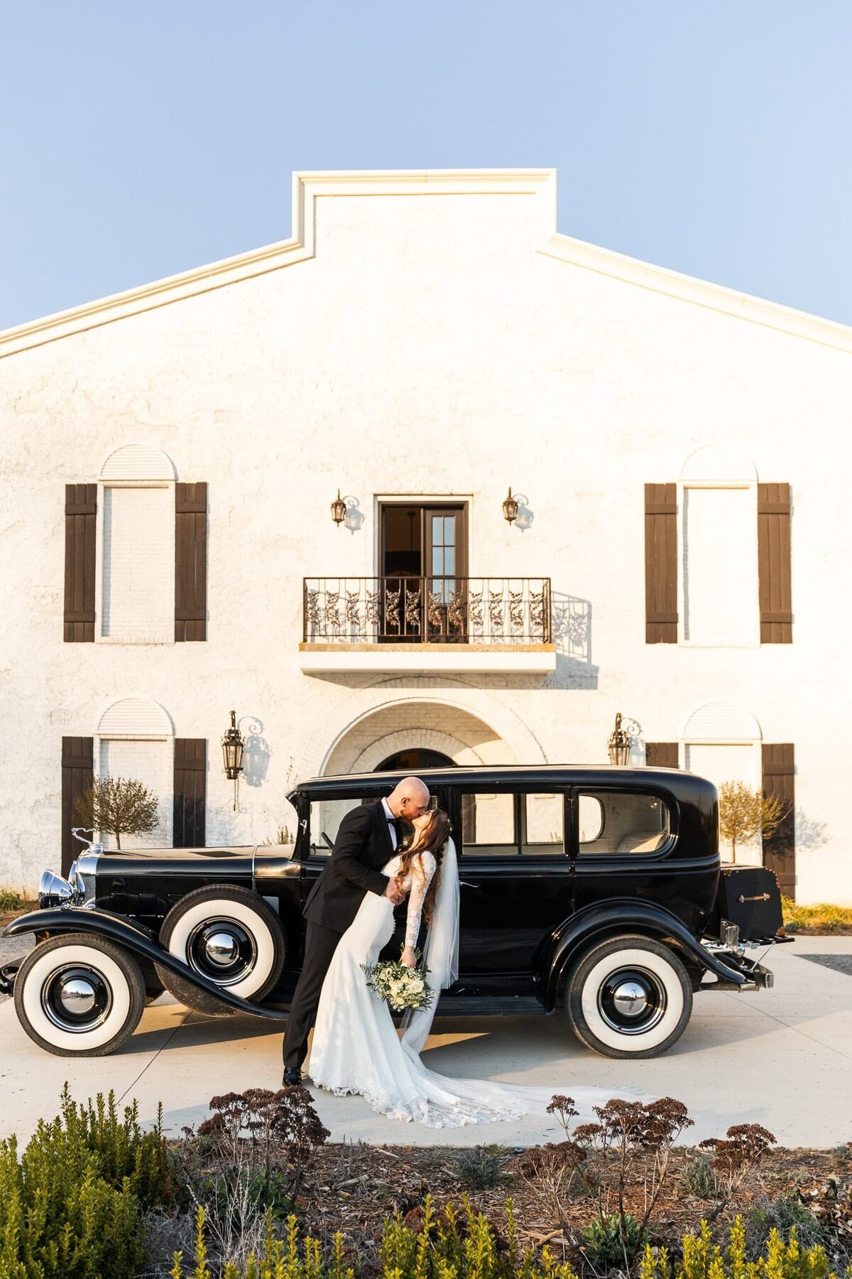 A newlywed couple shares a timeless kiss beside a vintage black car parked in front of the European-inspired white at Howe Farms in Chattanooga. The groom, dressed in a black tuxedo, dips his bride, who wears a long-sleeved lace gown, as the golden sunset bathes them in light