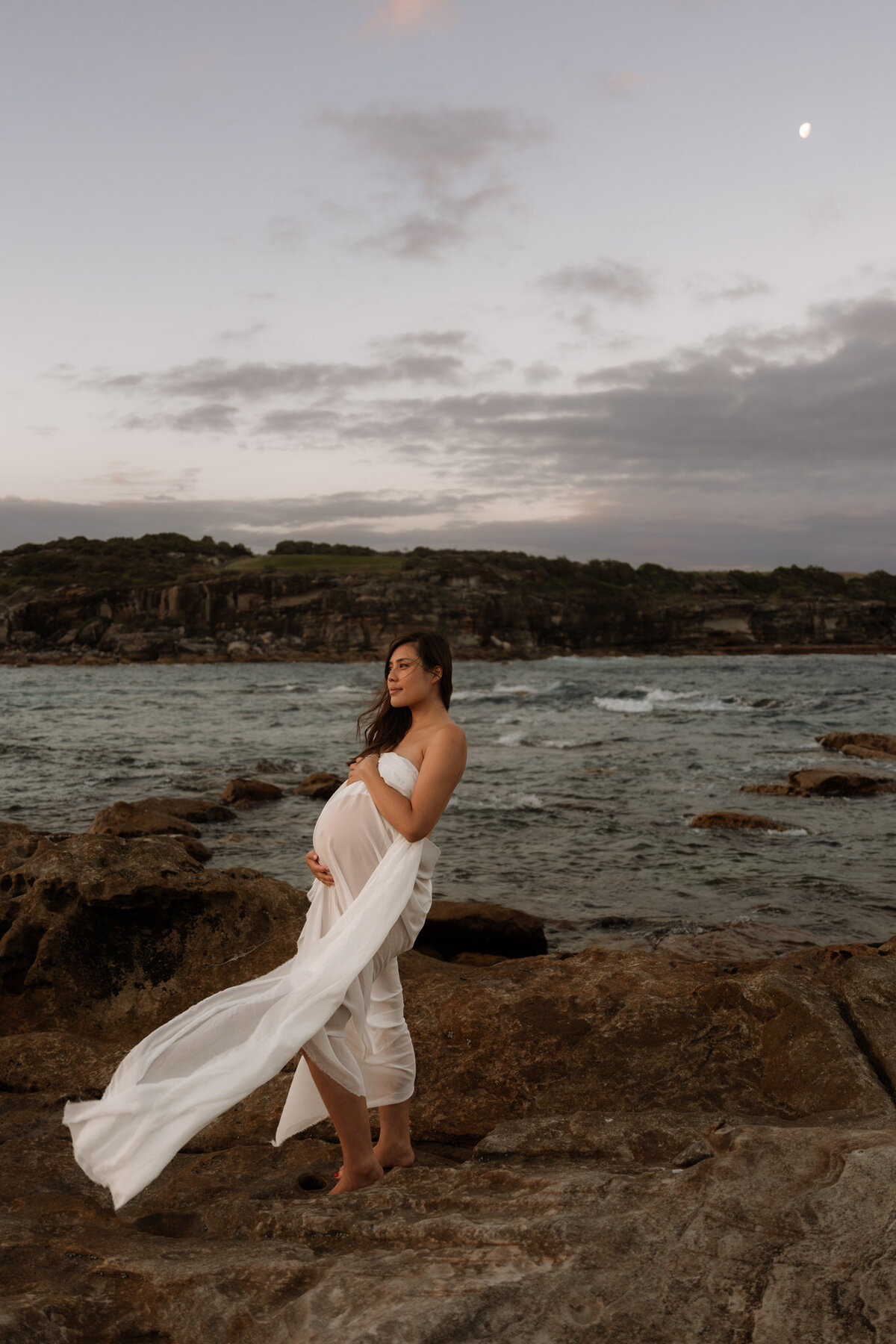 Pregnant woman in a flowing white sheet standing on coastal rocks at sunset during a maternity photography session in Sydney.