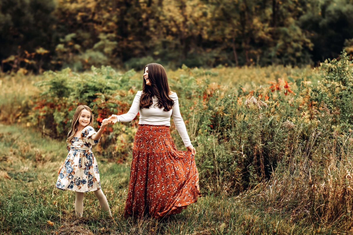 A woman and a young girl joyfully walk through a lush, green field. The woman wears a white top and a red patterned skirt, while the girl wears a floral dress. They hold hands, surrounded by tall grass and a backdrop of trees.