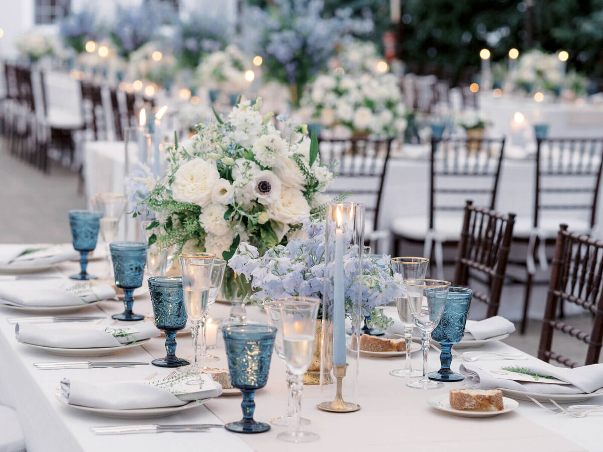 An elegant wedding dining table with a white flower centerpiece, cutleries, blue wine glasses, and candles. Image by Jenny Fu Studio