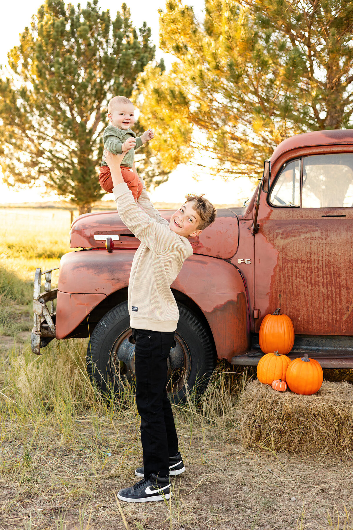 Preteen boy holds baby brother up in the air and they both smile.